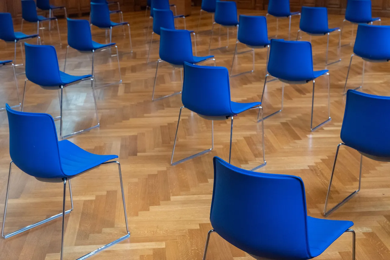 Ahşap parke zeminde sıralanmış mavi sandalyeler; genel kurul toplantısı için hazırlanmış salon